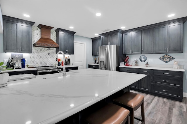 kitchen featuring light hardwood / wood-style floors, premium range hood, light stone counters, a breakfast bar area, and stainless steel fridge