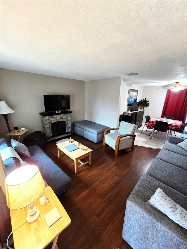 living room featuring a textured ceiling, ceiling fan, a stone fireplace, and dark wood-type flooring