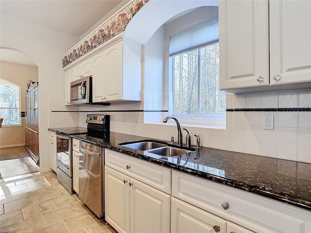 kitchen featuring a sink, tasteful backsplash, dark stone counters, arched walkways, and appliances with stainless steel finishes