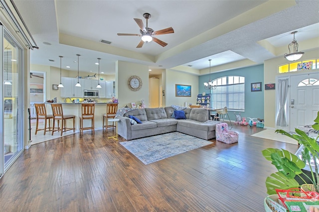 living room featuring ceiling fan with notable chandelier, a tray ceiling, and dark wood-type flooring