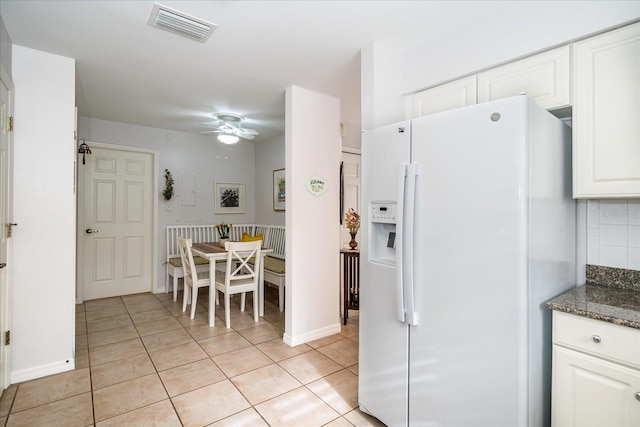 kitchen featuring ceiling fan, white fridge with ice dispenser, light tile patterned floors, dark stone counters, and white cabinets