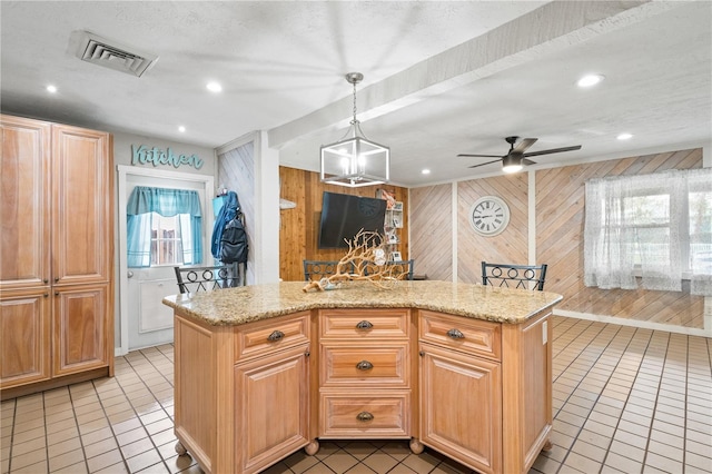 kitchen with pendant lighting, wood walls, a center island with sink, ceiling fan, and light stone counters