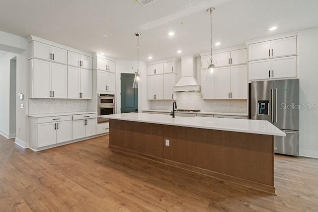 kitchen featuring premium range hood, appliances with stainless steel finishes, pendant lighting, a kitchen island with sink, and white cabinets