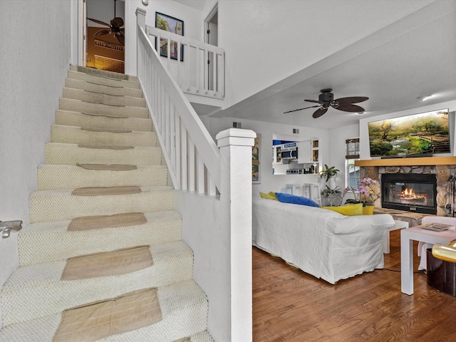 staircase with ceiling fan, wood-type flooring, and a stone fireplace