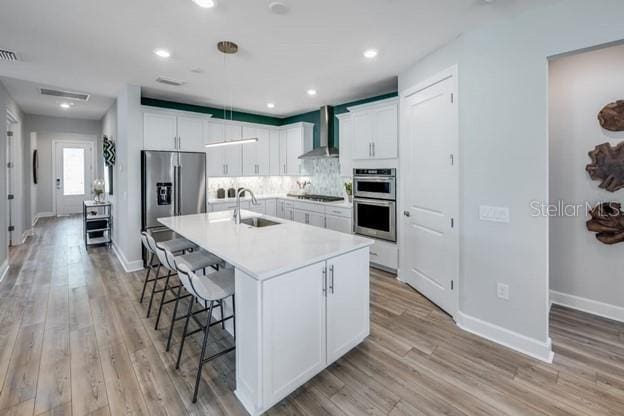 kitchen featuring sink, wall chimney exhaust hood, an island with sink, appliances with stainless steel finishes, and white cabinetry