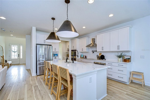 kitchen with white cabinetry, wall chimney range hood, stainless steel refrigerator with ice dispenser, a breakfast bar area, and a kitchen island with sink