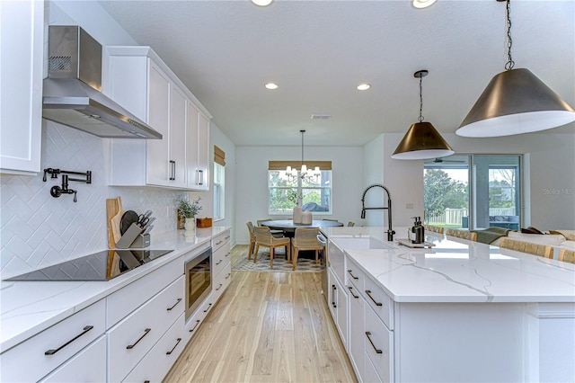 kitchen with light stone countertops, white cabinetry, wall chimney exhaust hood, an island with sink, and pendant lighting