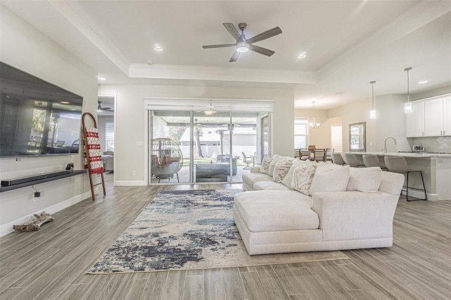 living room with ceiling fan with notable chandelier, crown molding, sink, and a tray ceiling