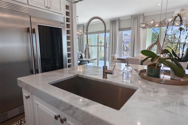 kitchen with light stone counters, a sink, hanging light fixtures, white cabinetry, and stainless steel built in refrigerator