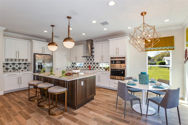 kitchen with pendant lighting, a kitchen island with sink, wall chimney exhaust hood, white cabinetry, and stainless steel appliances