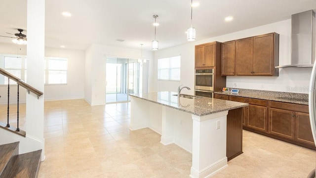 kitchen featuring a center island with sink, ceiling fan, hanging light fixtures, wall chimney exhaust hood, and sink