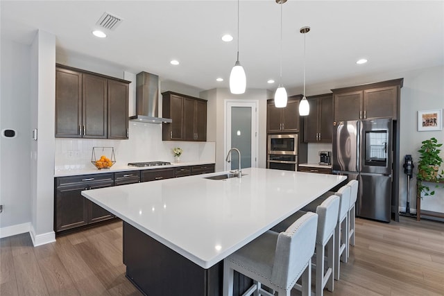 kitchen featuring wall chimney range hood, pendant lighting, sink, appliances with stainless steel finishes, and an island with sink