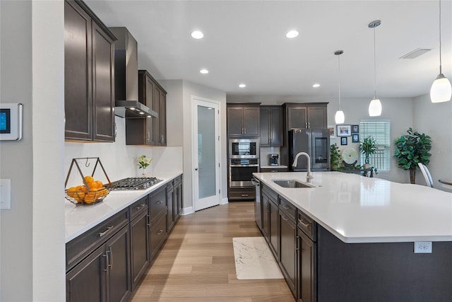 kitchen featuring pendant lighting, appliances with stainless steel finishes, wall chimney range hood, an island with sink, and sink