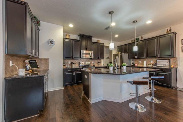 kitchen with dark brown cabinets, stainless steel appliances, dark wood-type flooring, pendant lighting, and a center island with sink