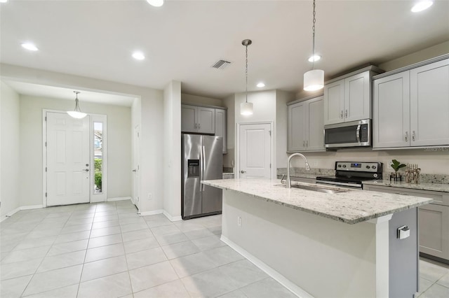 kitchen with pendant lighting, appliances with stainless steel finishes, gray cabinetry, light stone countertops, and a kitchen island with sink