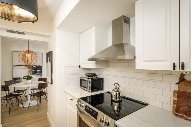 kitchen with white cabinets, appliances with stainless steel finishes, hanging light fixtures, and wall chimney range hood