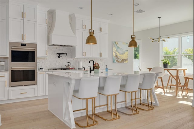 kitchen with an island with sink, stainless steel double oven, custom range hood, pendant lighting, and white cabinets