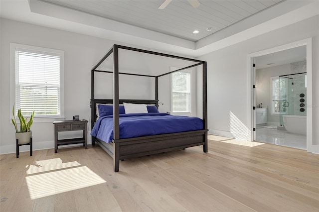 bedroom with light wood-type flooring, ceiling fan, a raised ceiling, and ensuite bath