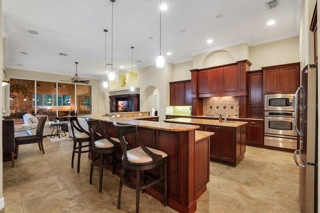 kitchen featuring ceiling fan, stainless steel appliances, backsplash, decorative light fixtures, and a center island with sink