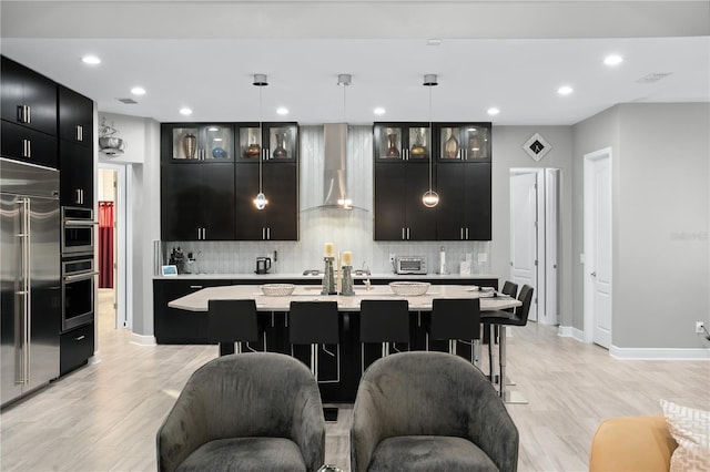 kitchen with high end fridge, an island with sink, wall chimney exhaust hood, and pendant lighting