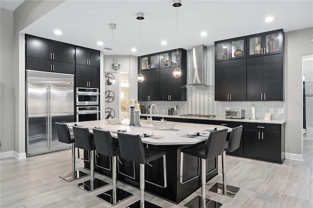 kitchen featuring appliances with stainless steel finishes, hanging light fixtures, an island with sink, a kitchen bar, and wall chimney range hood