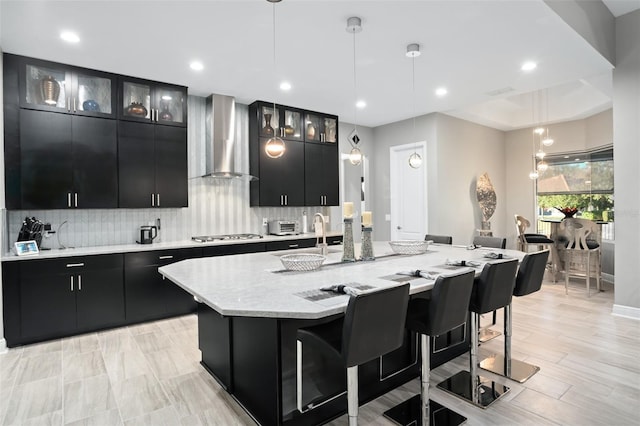 kitchen with a breakfast bar, pendant lighting, a kitchen island with sink, and wall chimney exhaust hood