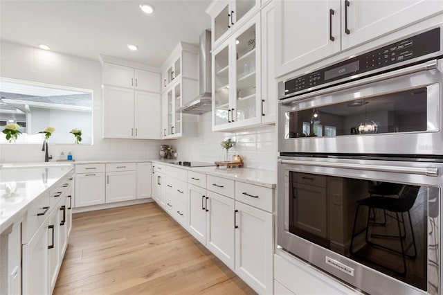 kitchen with white cabinets, light stone countertops, stainless steel double oven, and wall chimney range hood