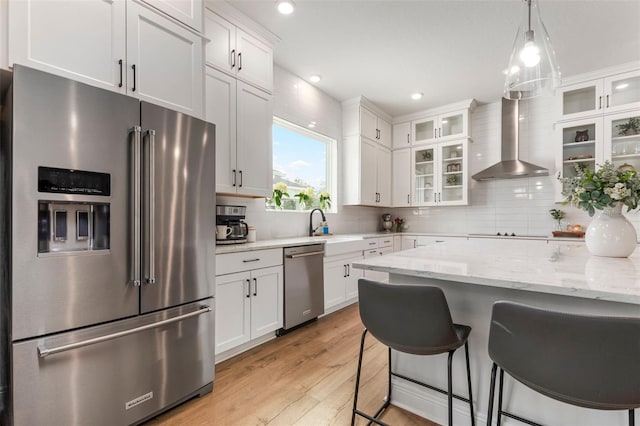 kitchen with appliances with stainless steel finishes, wall chimney range hood, decorative light fixtures, white cabinets, and light hardwood / wood-style floors