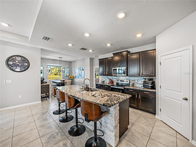 kitchen with dark stone countertops, a breakfast bar area, dark brown cabinets, a center island with sink, and appliances with stainless steel finishes