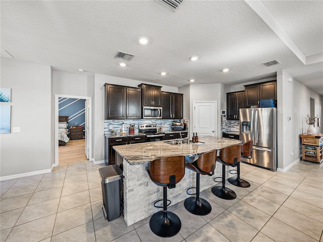 kitchen featuring an island with sink, decorative backsplash, a breakfast bar area, appliances with stainless steel finishes, and light tile patterned flooring