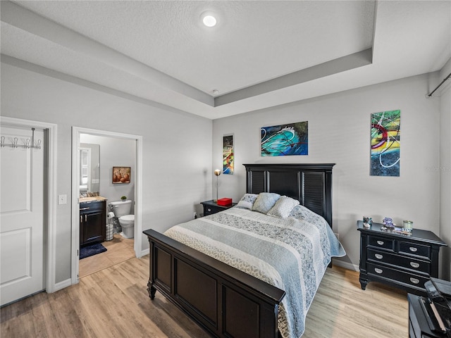 bedroom featuring light wood-type flooring, a tray ceiling, a textured ceiling, and connected bathroom