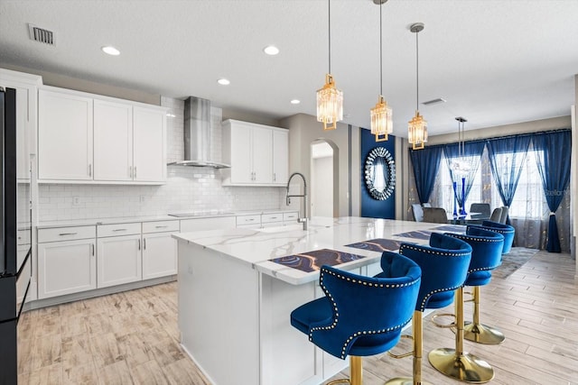 kitchen featuring decorative light fixtures, a kitchen bar, white cabinetry, wall chimney range hood, and a kitchen island with sink