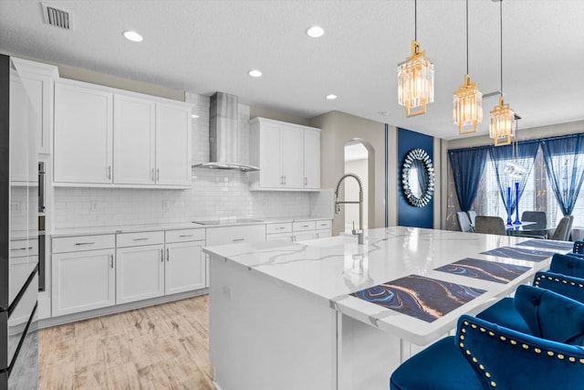 kitchen featuring light stone countertops, white cabinets, wall chimney exhaust hood, decorative light fixtures, and a kitchen island with sink