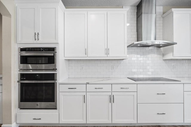 kitchen featuring tasteful backsplash, wall chimney range hood, stainless steel double oven, white cabinets, and black electric cooktop