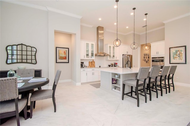 kitchen with white cabinetry, wall chimney range hood, stainless steel appliances, hanging light fixtures, and a kitchen island with sink