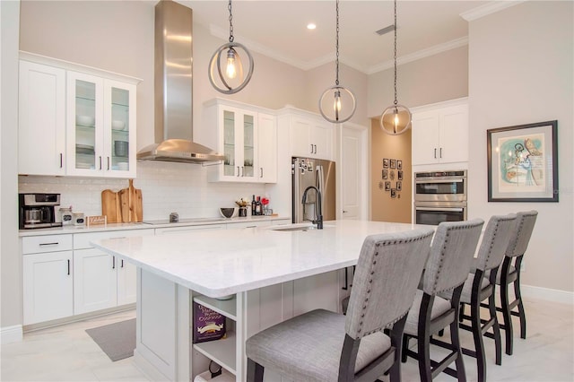 kitchen featuring an island with sink, stainless steel appliances, tasteful backsplash, wall chimney range hood, and white cabinets