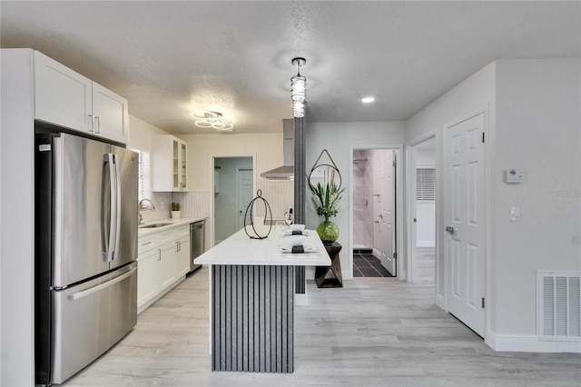 kitchen featuring appliances with stainless steel finishes, decorative light fixtures, white cabinets, wall chimney range hood, and a center island with sink