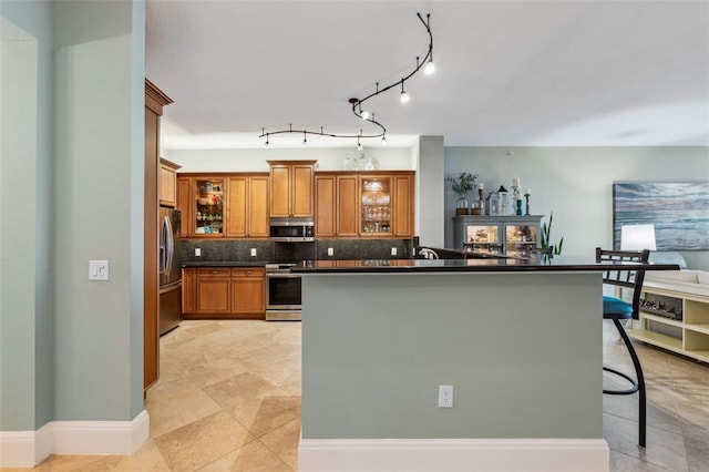 kitchen featuring brown cabinetry, dark countertops, glass insert cabinets, stainless steel appliances, and backsplash