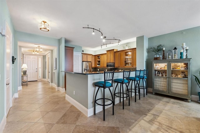 kitchen featuring a peninsula, appliances with stainless steel finishes, brown cabinetry, dark countertops, and glass insert cabinets