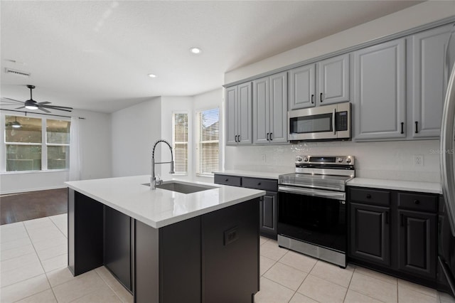 kitchen with appliances with stainless steel finishes, sink, light tile patterned floors, gray cabinets, and an island with sink