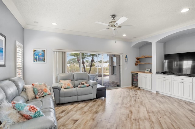 living room featuring a textured ceiling, beverage cooler, light wood-type flooring, ceiling fan, and crown molding