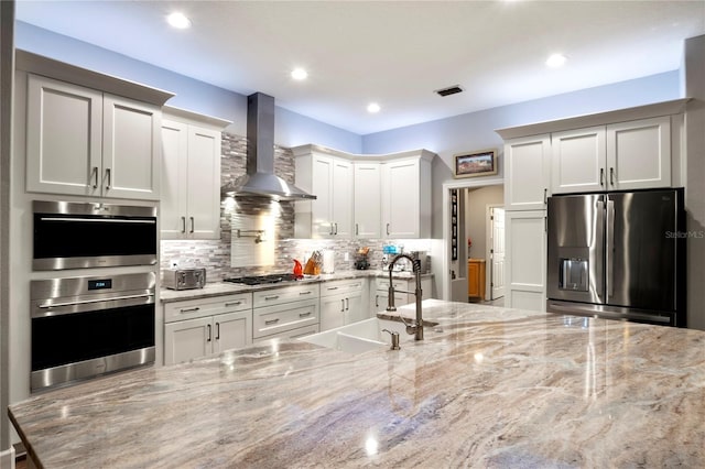 kitchen featuring stainless steel appliances, light stone countertops, wall chimney exhaust hood, white cabinets, and sink