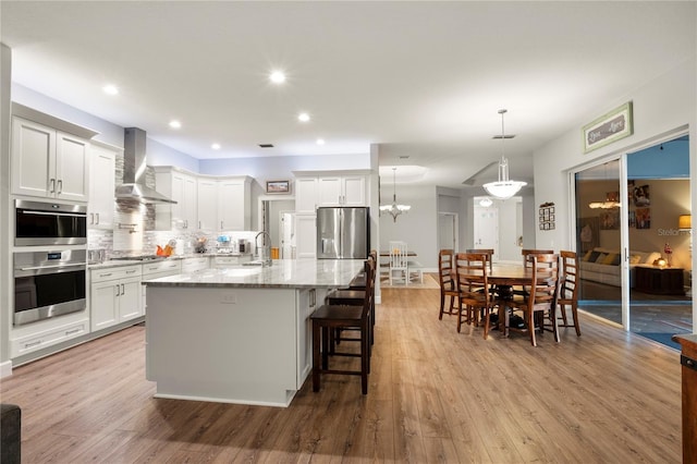 kitchen with backsplash, a center island with sink, white cabinetry, stainless steel appliances, and wall chimney exhaust hood