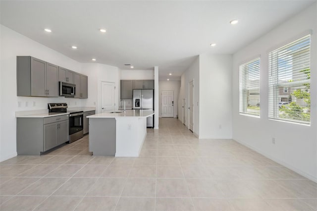 kitchen with stainless steel appliances, sink, light tile patterned floors, gray cabinets, and a kitchen island with sink