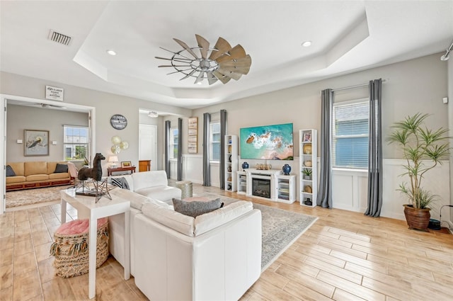 living room featuring a raised ceiling, ceiling fan, a wealth of natural light, and light wood-type flooring