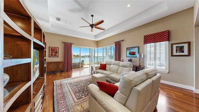 living room with a tray ceiling, ceiling fan, a water view, and dark wood-type flooring