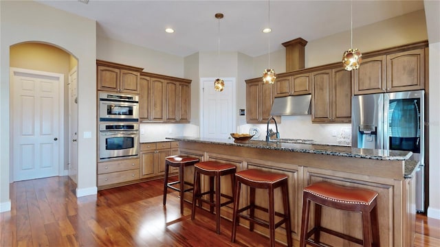 kitchen with hanging light fixtures, stainless steel appliances, dark stone countertops, an island with sink, and a kitchen bar