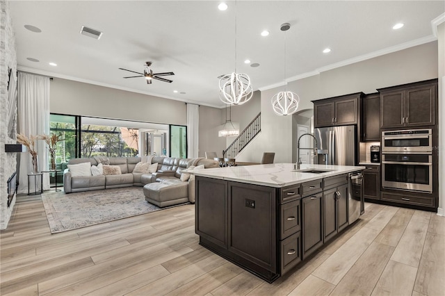 kitchen featuring appliances with stainless steel finishes, hanging light fixtures, an island with sink, dark brown cabinetry, and ceiling fan with notable chandelier