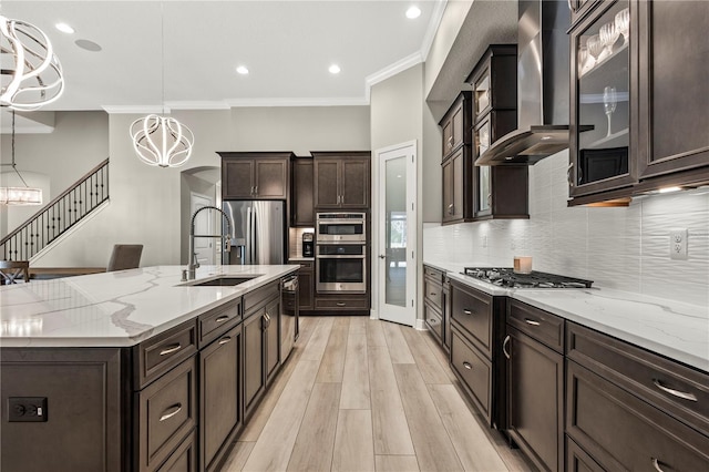 kitchen featuring wall chimney exhaust hood, an inviting chandelier, a kitchen island with sink, appliances with stainless steel finishes, and ornamental molding