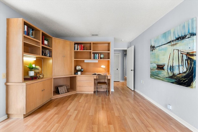 office featuring a textured ceiling, visible vents, baseboards, light wood finished floors, and built in desk